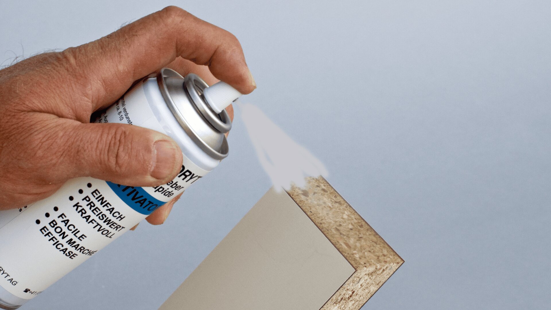 Eine Hand sprüht Klebstoff aus einer Spraydose auf die Kante einer Spanplatte vor einem unbedruckten Hintergrund.