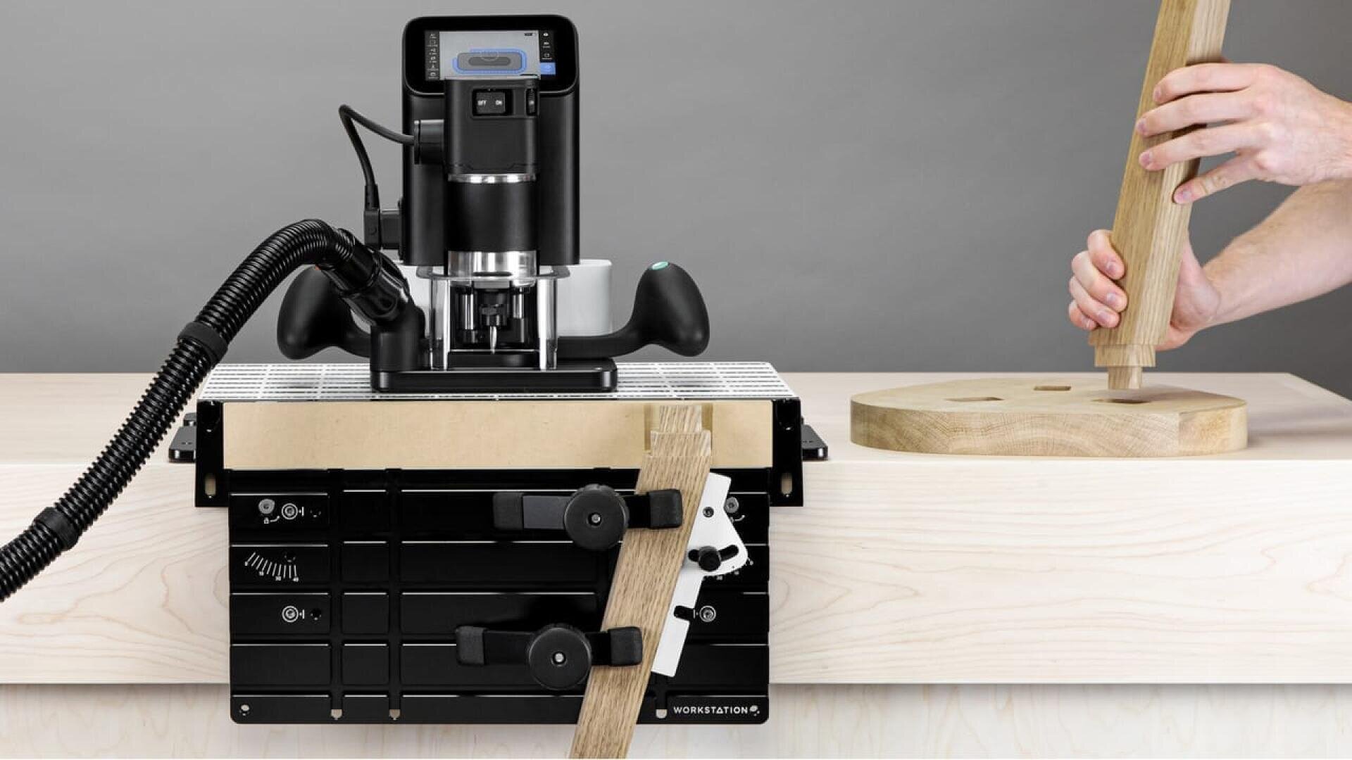 Une personne utilise un outil de travail du bois avec une défonceuse noire et un accessoire sur un établi en bois léger, tenant une pièce en bois tout en assemblant ou en fabriquant un autre article en bois.
