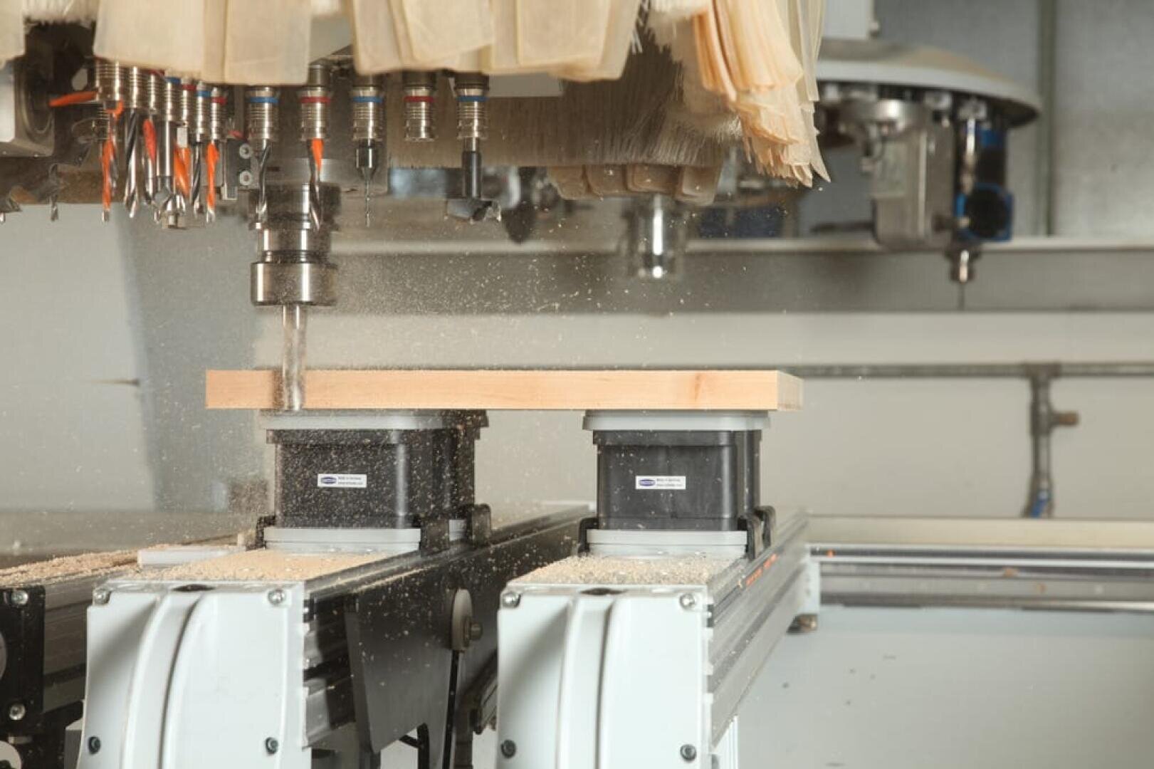 Eine CNC-Maschine bohrt in eine Holzplatte, die von Absaugklammern gehalten wird, wobei Holzspäne und Staub im Arbeitsbereich verstreut werden. Oben sind mehrere Werkzeugköpfe und eine Staubabsaugbürste zu sehen.