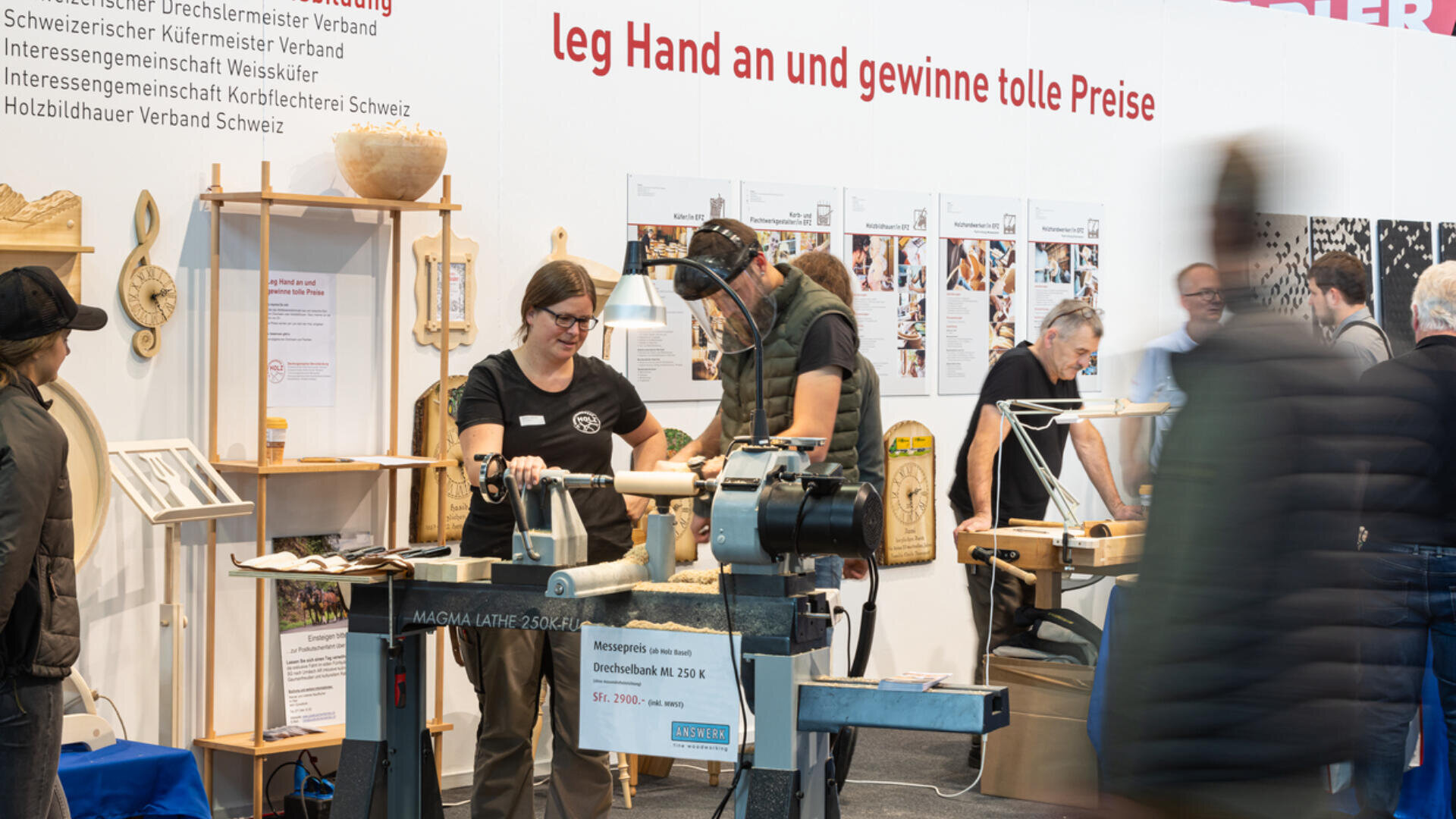 Menschen nehmen an einer Holzbearbeitungsveranstaltung teil und benutzen eine Drehmaschine. Eine Frau erklärt den Prozess, während ein Mann an der Maschine arbeitet. Andere beobachten oder interagieren in dem belebten Ausstellungsraum. Informationsplakate bedecken die Wand.