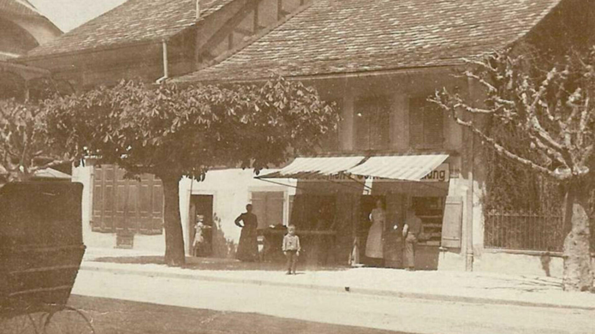 Sepiafarbenes Foto von Menschen, die vor einem Geschäft mit Markise stehen, in einer von beschnittenen Bäumen und alten Gebäuden gesäumten Straße; links ist teilweise eine Pferdekutsche zu sehen.