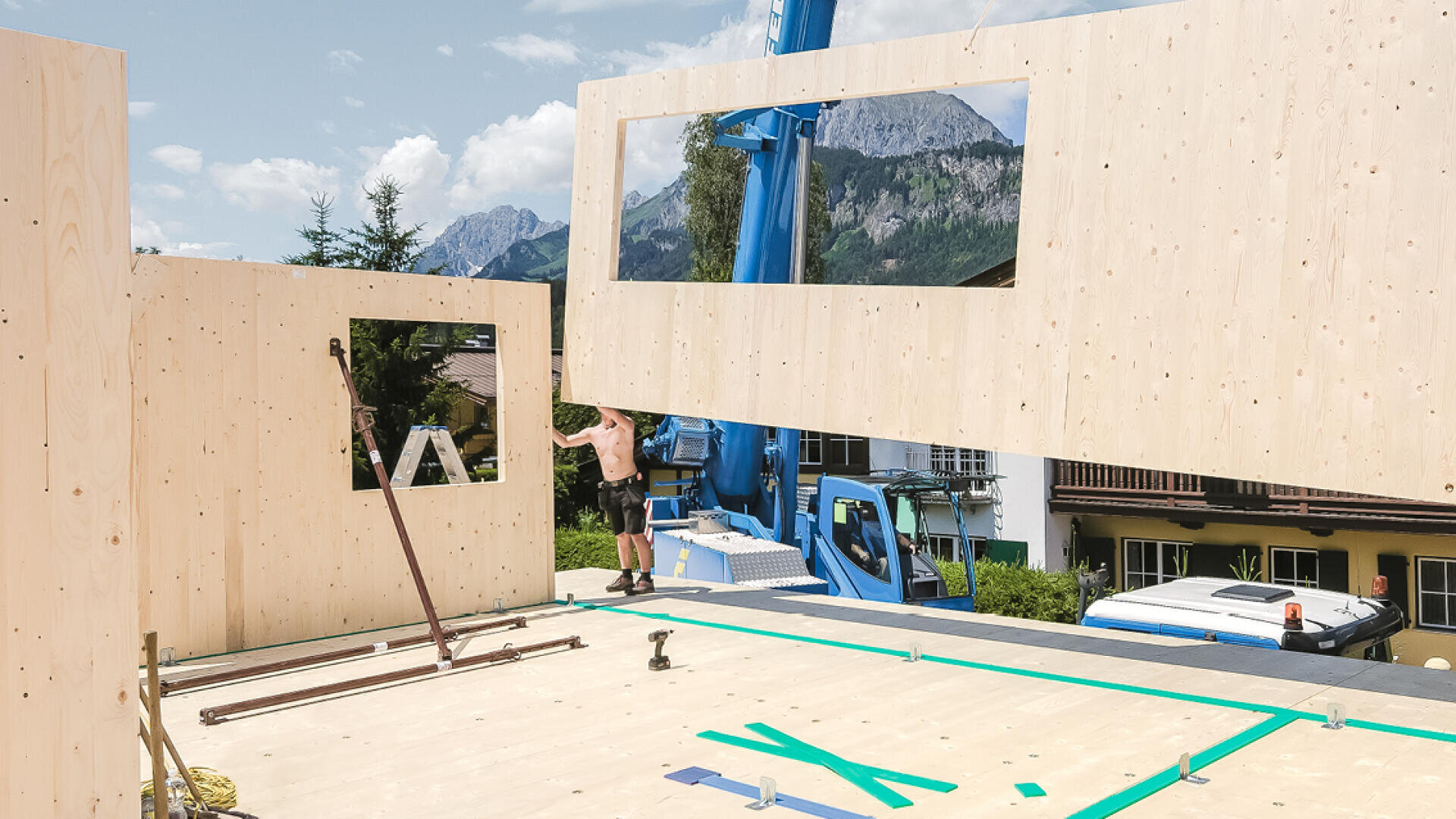 Un ouvrier de la construction se tient sur un chantier en bois tandis qu'une grue soulève un grand panneau mural en bois avec des découpes de fenêtres. Des montagnes et des arbres sont visibles à l'arrière-plan sous un ciel partiellement nuageux.
