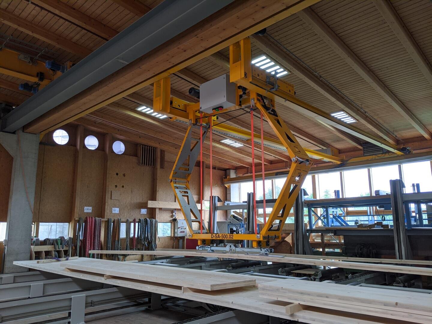 Ein gelber industrieller Heberoboter wird in einer Tischlerei auf Schienen über gestapelten Holzbrettern montiert. Der Arbeitsbereich umfasst Holzbalken, Werkzeuge und Fenster, die für natürliches Licht sorgen.