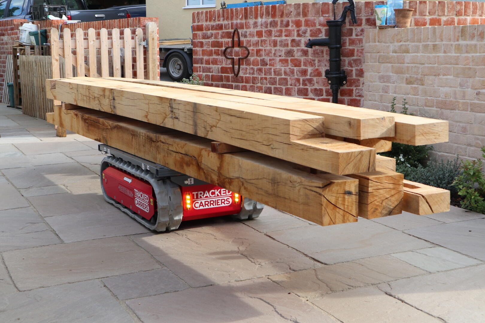 Ein kleiner roter Tragroboter transportiert mehrere große Holzbalken über eine gepflasterte Terrasse mit Ziegelmauern und Zäunen im Hintergrund.