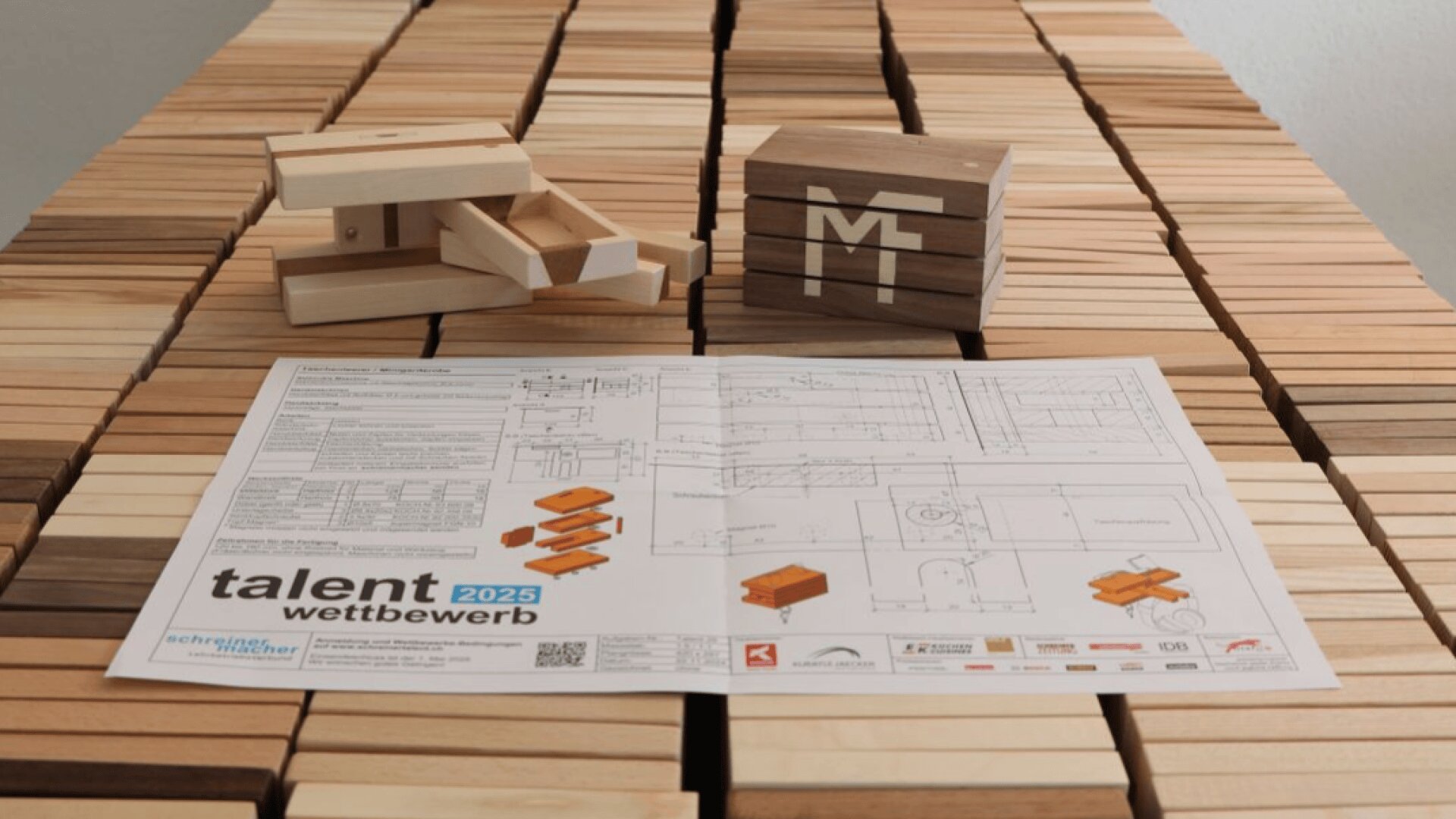Une feuille avec des dessins techniques et le talent wettbewerb 2025 est posée sur une table recouverte de blocs de bois. Deux maquettes en bois et un cube portant les initiales de MT sont également posés sur la table.
