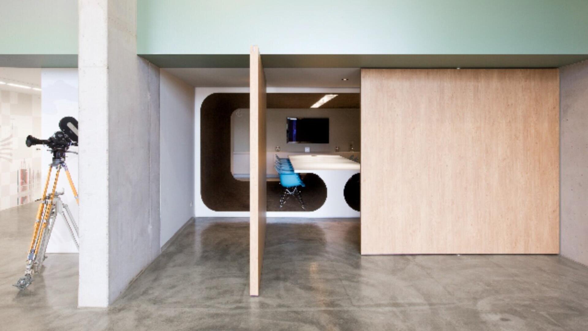 Un espace de bureau moderne avec des sols en béton poli, une porte coulissante en bois clair partiellement ouverte pour révéler un espace de réunion futuriste avec des découpes murales géométriques et une chaise bleue. Un appareil photo vintage est posé sur un trépied à gauche.