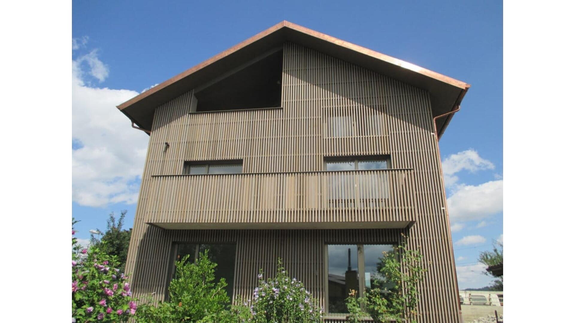 Une maison moderne de deux étages avec des lattes de bois verticales couvrant la façade, de grandes fenêtres, un toit en pente et un jardin avec des plantes vertes et des buissons fleuris devant, sous un ciel bleu avec des nuages épars.