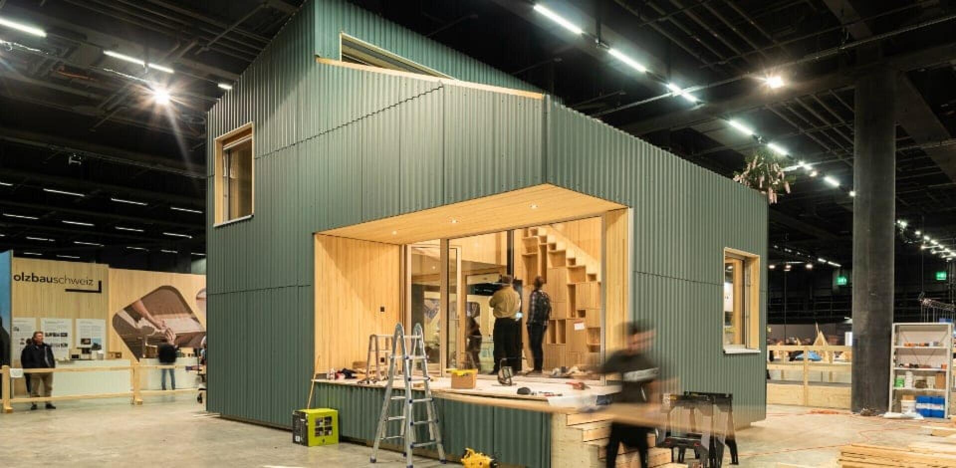 Ein modernes, vorgefertigtes Häuschen mit grüner Wellblechverkleidung und großen Fenstern mit Holzrahmen ist in einem Innenraum ausgestellt. Um das Haus herum wird gearbeitet, und in der Nähe liegen Werkzeuge und Materialien unter hellen Deckenleuchten verstreut.