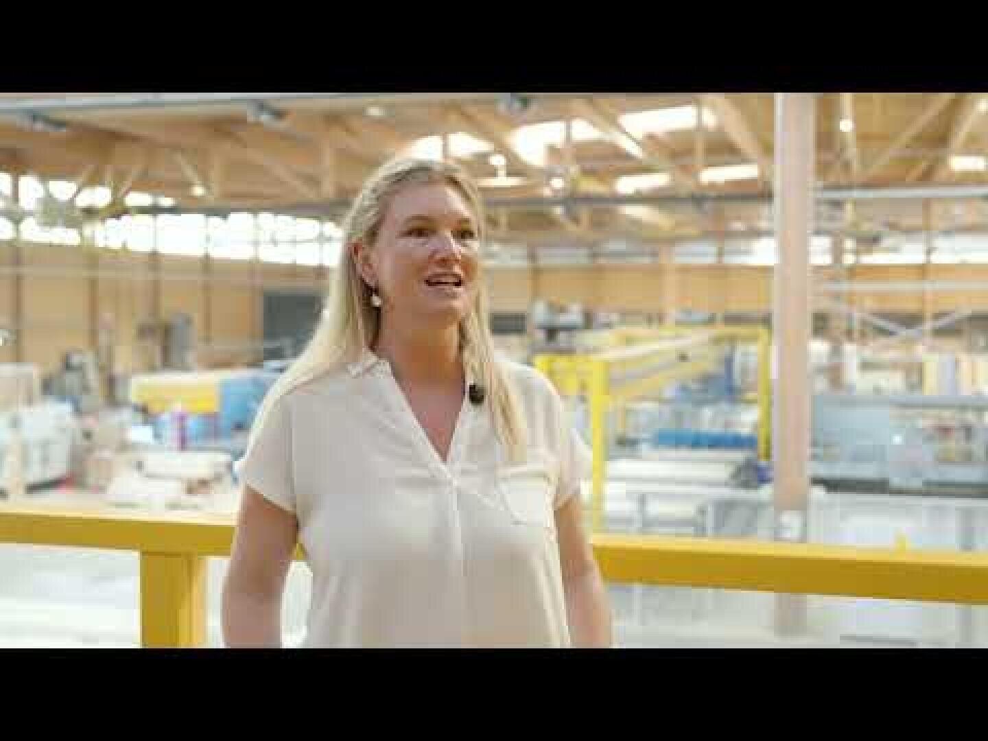 Eine Frau mit langen blonden Haaren und weißer Bluse steht in einem Innenraum und lächelt. Sie steht vor einem gelben Geländer mit Blick auf ein großes Industrielager mit verschiedenen Maschinen und natürlichem Licht.