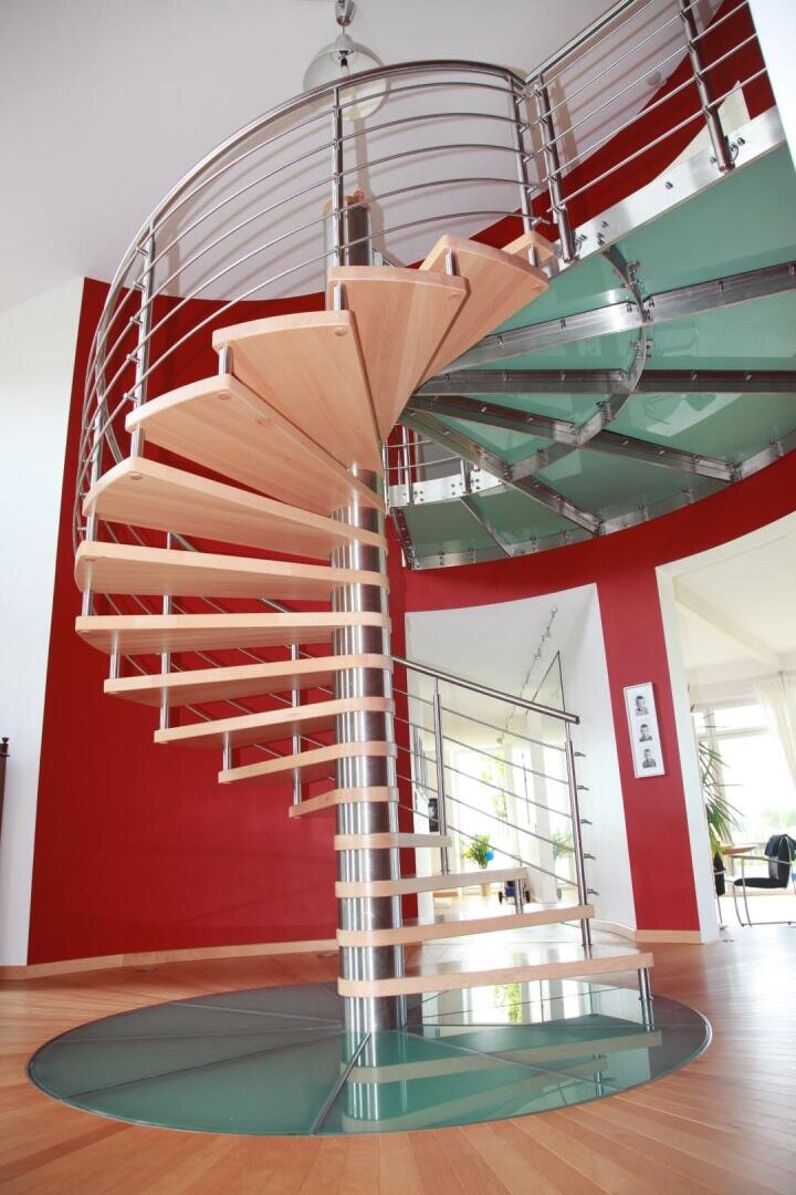 Eine moderne Spindeltreppe mit Holzstufen und Metallgeländer steht auf einem runden Glassockel in einem hellen Raum mit roten Wänden und Holzfußböden. Durch große Fenster im Hintergrund fällt natürliches Licht ein.