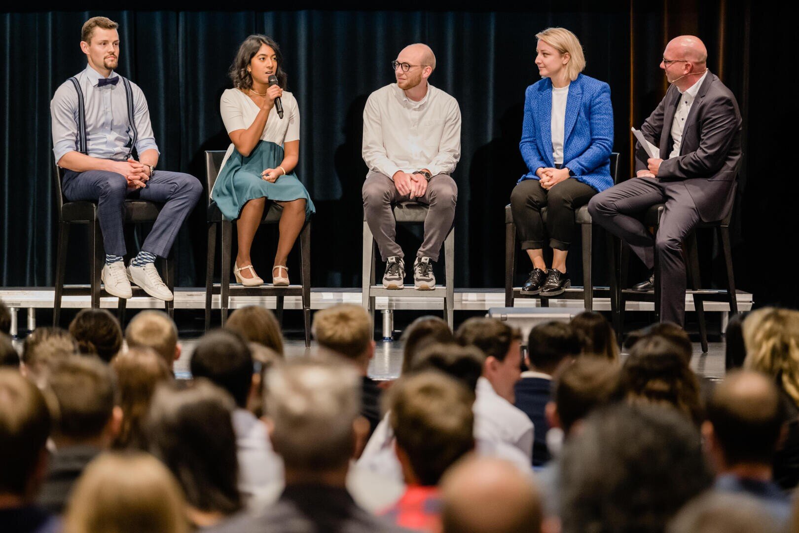 Cinq personnes sont assises sur une scène pendant une discussion de podium devant un public. Une personne tient un micro et parle pendant que les autres écoutent. Tous sont vêtus d'une tenue professionnelle décontractée.