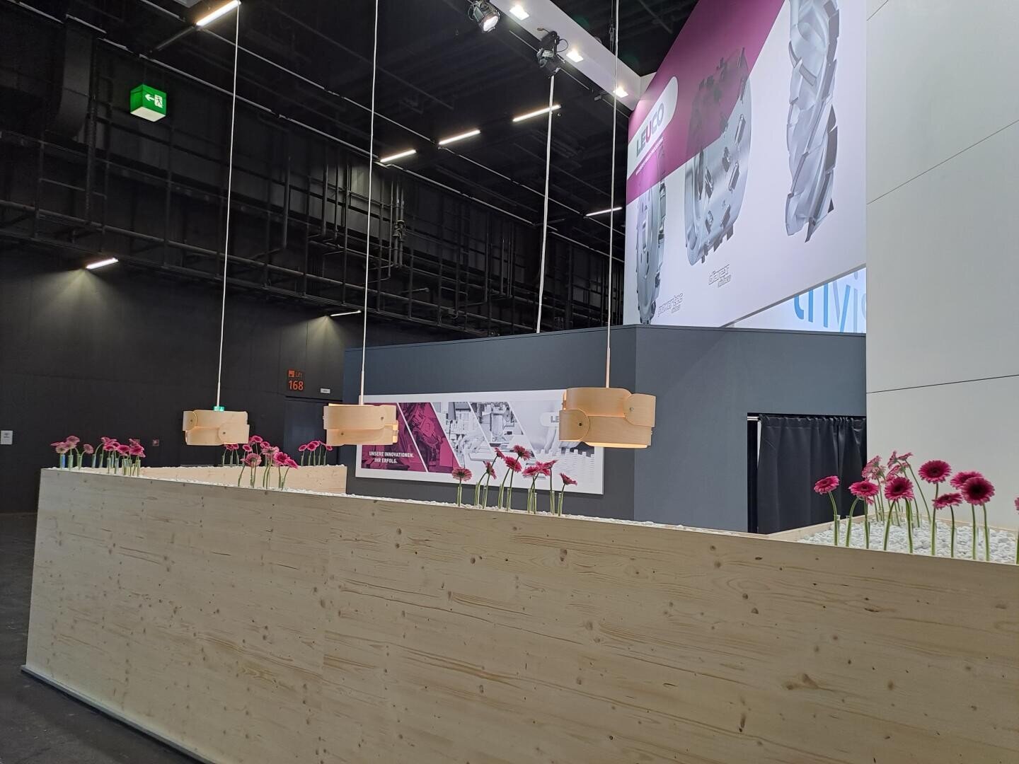 Ein moderner Innenraum mit einer langen Holztheke mit gleichmäßig verteilten rosa Blumen in Vasen, darüber hängenden Pendelleuchten und dunklen Decken im Industriestil. Im Hintergrund befinden sich große Poster und eine graue Trennwand.
