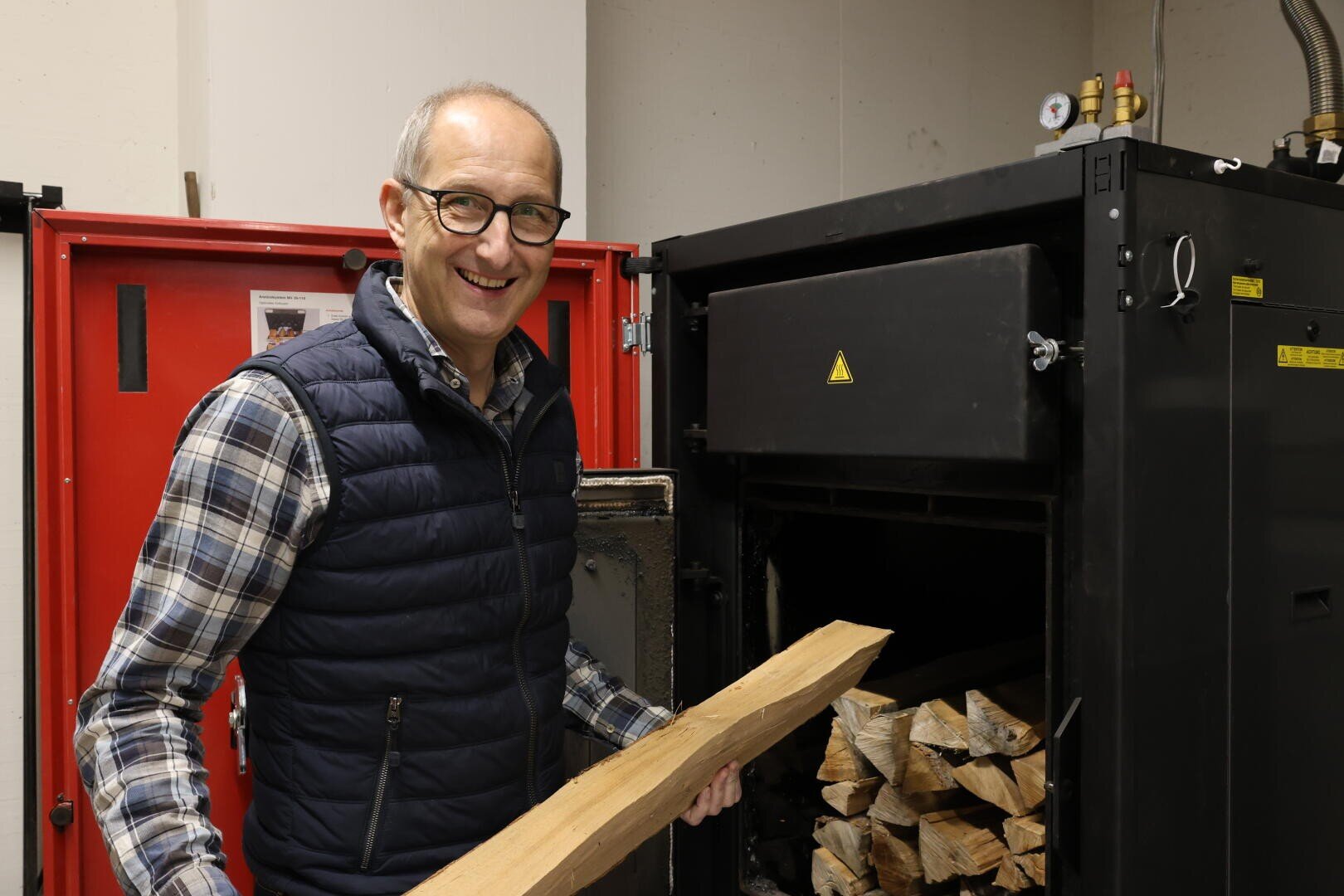 Ein lächelnder Mann mit Brille und kariertem Hemd hält in einem Innenbereich ein Stück Brennholz vor einem mit Holzscheiten gefüllten Holzofen.