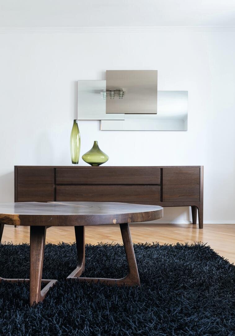 Ein modernes Wohnzimmer mit einem hölzernen Couchtisch auf einem schwarzen Zottelteppich, einem langen hölzernen Sideboard, zwei grünen Vasen und einem abstrakten rechteckigen Spiegel an einer weißen Wand.