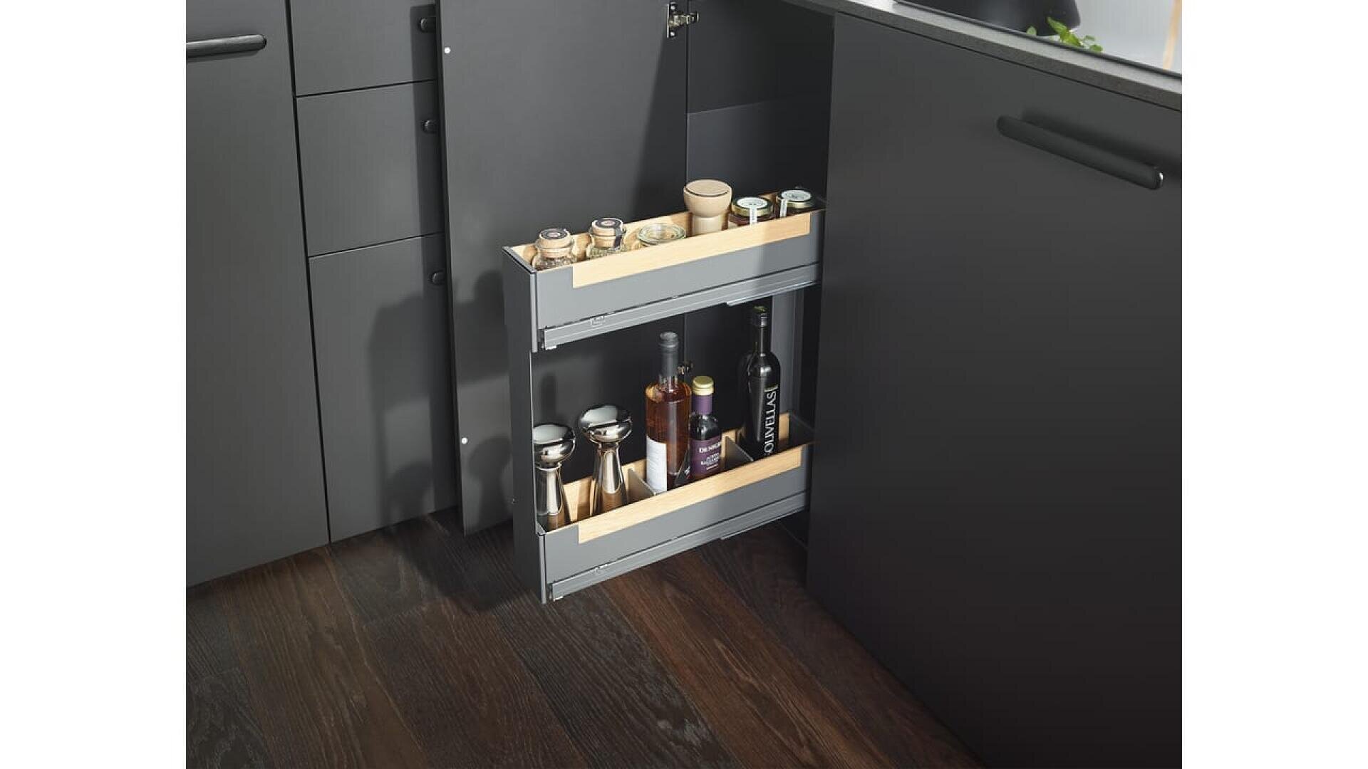 Un meuble de cuisine coulissant à deux étagères permet de ranger les bouteilles, les bocaux et les récipients dans un espace étroit entre les meubles sombres et l'évier.