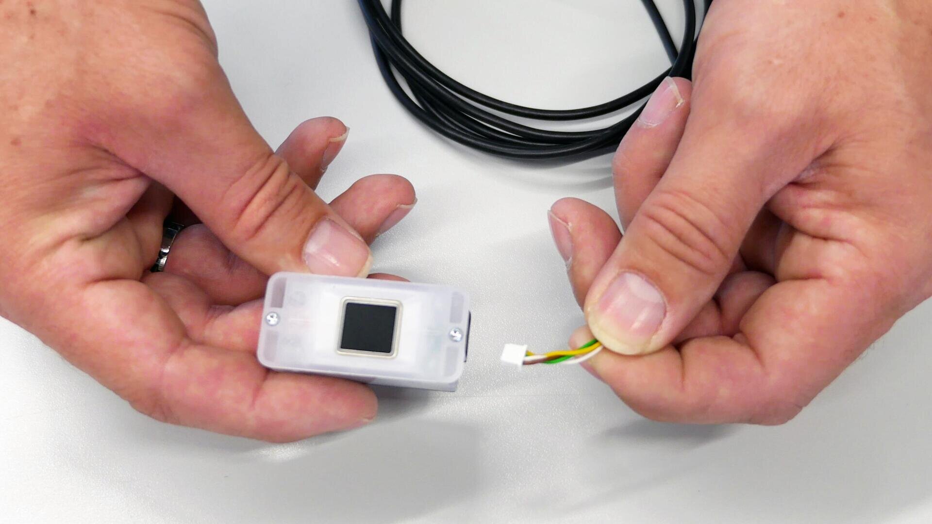 Deux mains tiennent des composants électroniques : l'une tient un petit capteur rectangulaire avec un carré noir, et l'autre tient un connecteur avec quatre fils colorés, au-dessus d'une surface blanche.