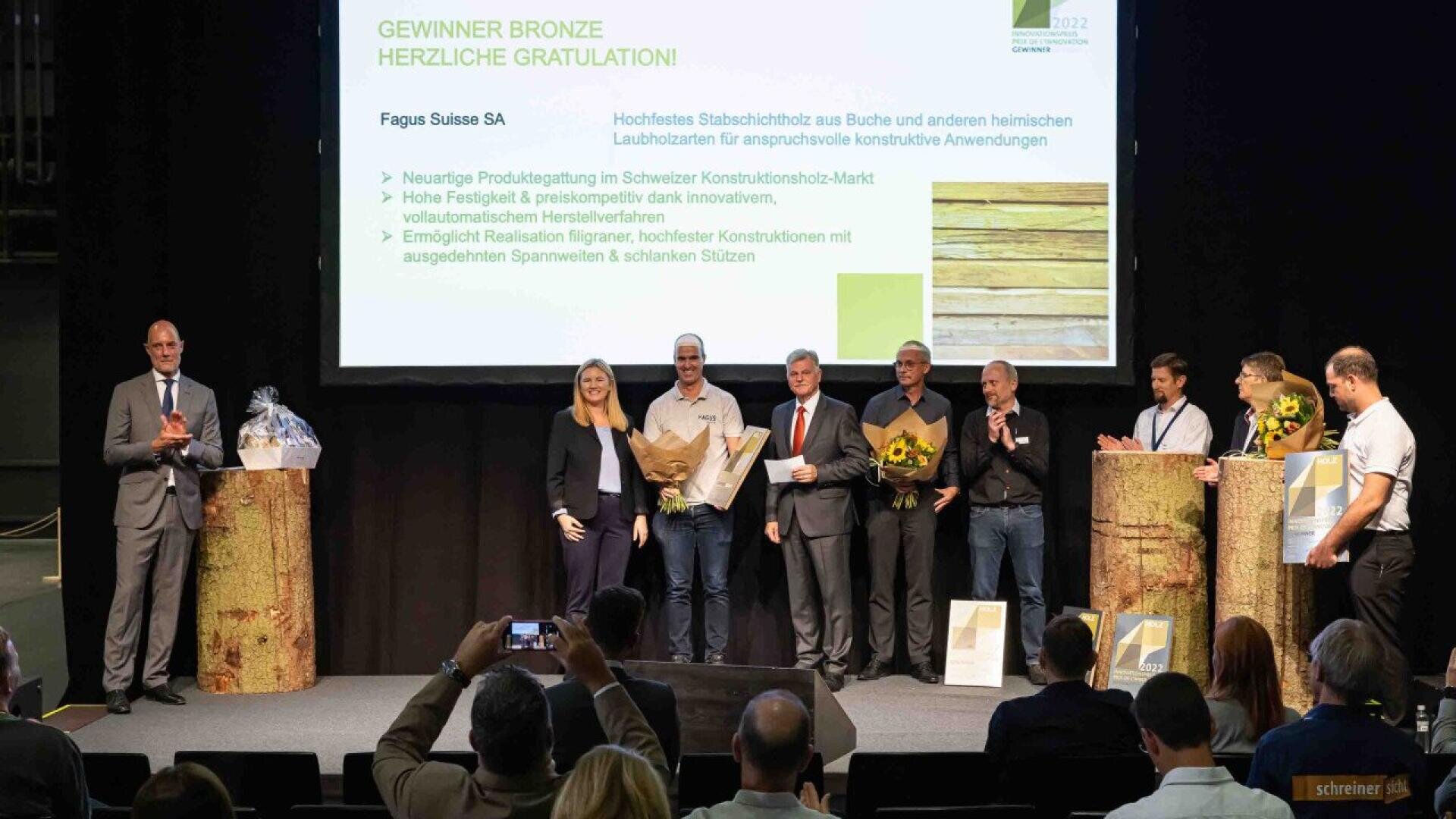 Un groupe de personnes sur scène lors d'une cérémonie de remise de prix, certaines tenant des fleurs et des certificats. Derrière eux, une diapositive de présentation annonce le lauréat de bronze, Fagus Suisse SA, avec un texte de félicitations en allemand.