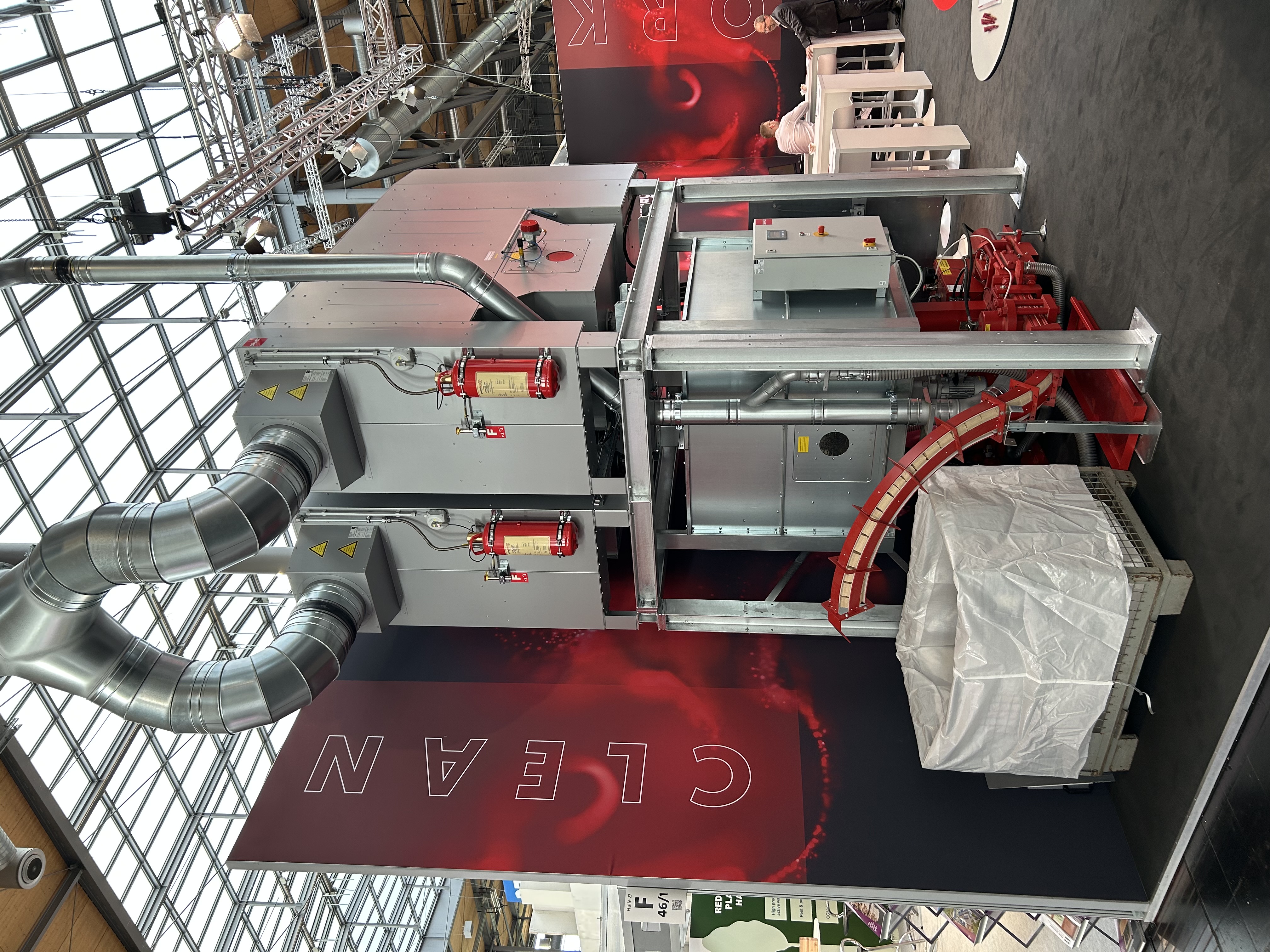 Große industrielle Luftfilteranlage mit Metallkanälen, Feuerlöschern und einem großen weißen Auffangbeutel darunter. Die Anlage ist auf einem Messestand mit dem Wort CLEAN auf einem rot-schwarzen Hintergrund zu sehen.