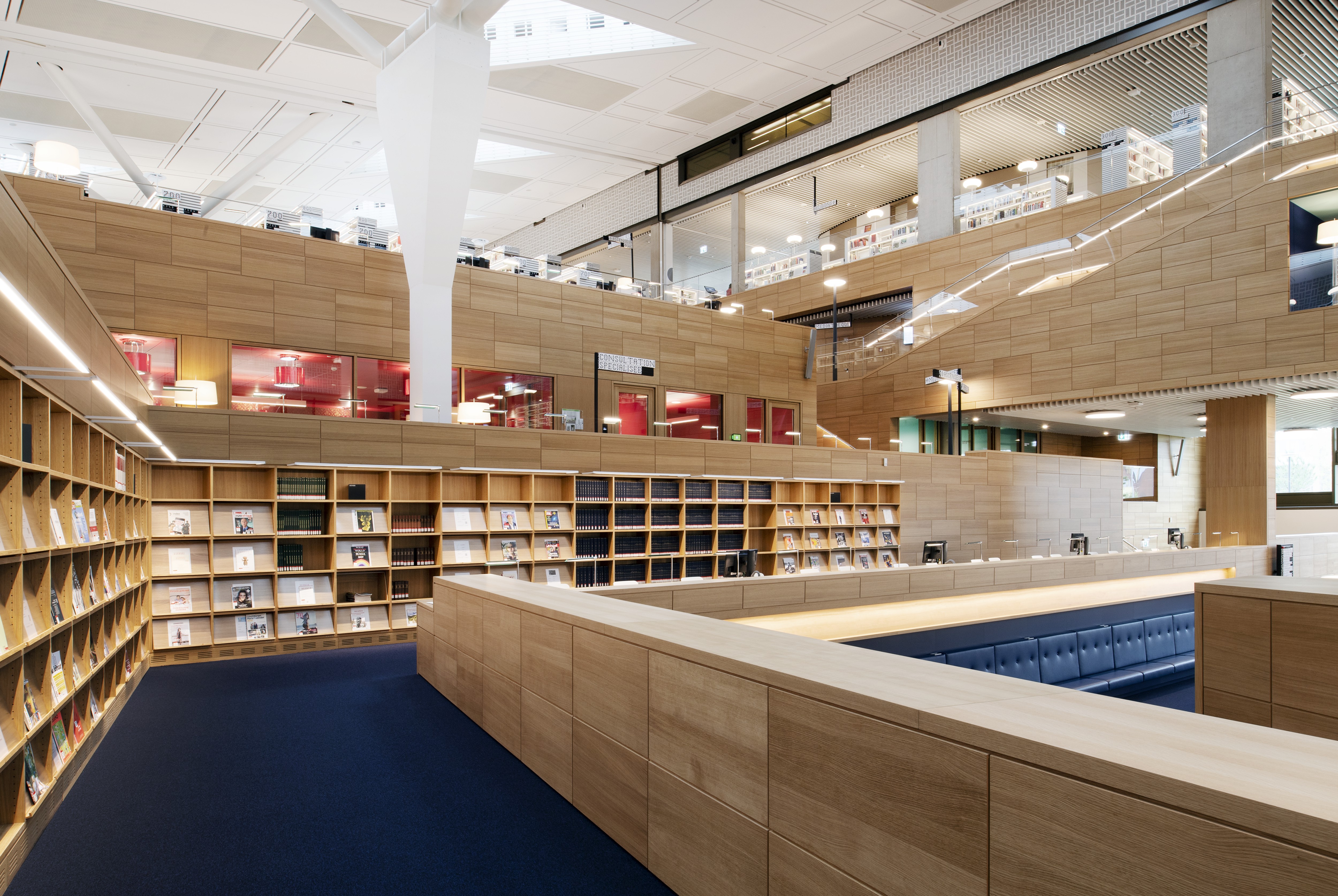 Am&eacute;nagement moderne de la biblioth&egrave;que avec des &eacute;tag&egrave;res en bois, un tapis bleu et des si&egrave;ges spacieux. De grandes fen&ecirc;tres et des balcons ouverts aux &eacute;tages sup&eacute;rieurs assurent la lumi&egrave;re naturelle et la vue sur d'autres biblioth&egrave;ques et espaces de travail.