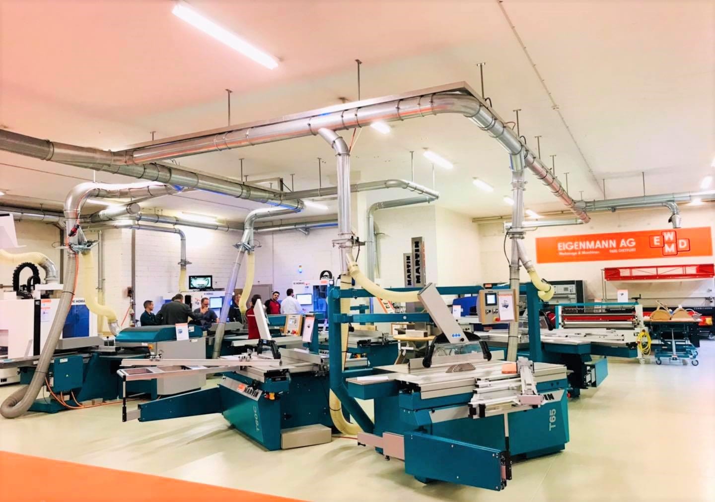 Eine helle, geräumige Fabrikhalle mit mehreren großen Industriemaschinen, Metallkanälen über dem Kopf und Menschen, die im Hintergrund arbeiten. Auf einem orangefarbenen Schild steht EIGENMANN AG. Die Umgebung wirkt sauber und organisiert.
