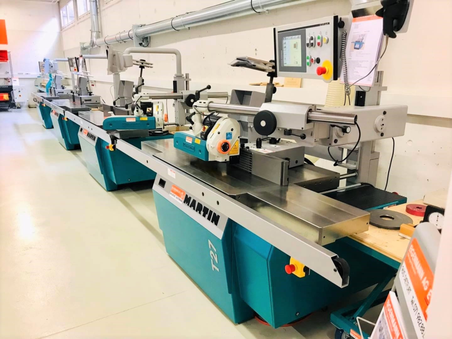 Eine Reihe von industriellen Holzbearbeitungsmaschinen mit digitalen Steuerungen in einer sauberen, gut beleuchteten Werkstatt. Die Maschinen sind türkisfarben und mit Martin beschriftet, und in der Nähe sind verschiedene Werkzeuge und Papiere zu sehen.