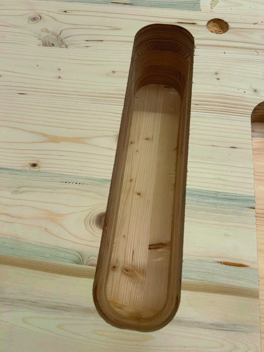 Gros plan sur une planche de bois dans laquelle une fente ovale allongée a été découpée, révélant le grain du bois à l'intérieur de la fente. La planche environnante présente des motifs naturels et des nœuds dans le bois.