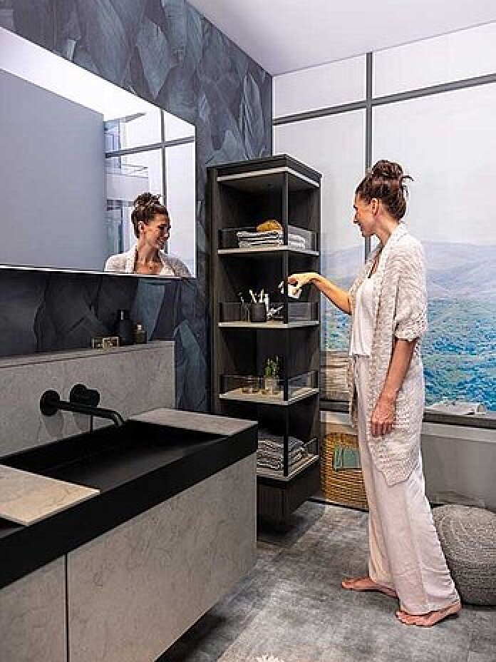 Eine Frau in leichter Loungewear steht in einem modernen Badezimmer und greift nach einem Gegenstand in einem hohen offenen Regal. Der Raum verfügt über einen großen Spiegel, ein schwarzes Waschbecken, einen Marmorarbeitsplatz und ein malerisches Wandgemälde an der Wand.