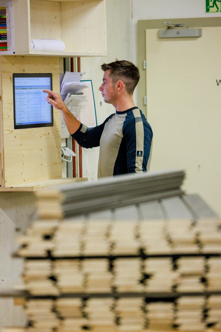 Un homme portant une chemise à manches longues utilise un écran tactile monté sur un mur en bois, avec une pile de planches en bois au premier plan.