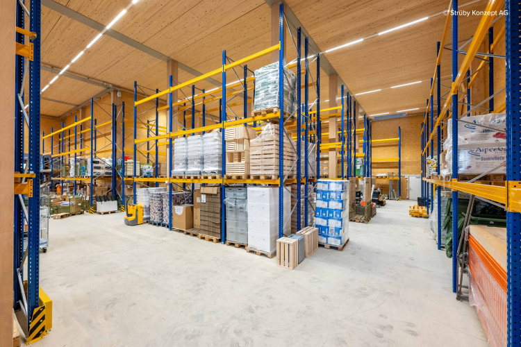 Ein geräumiges Lagerhaus mit hohen blauen und gelben Metallregalen, in denen Paletten mit Kisten, Paketen und Baumaterialien gelagert sind, alles ordentlich in Reihen unter einer hell beleuchteten Holzdecke angeordnet.
