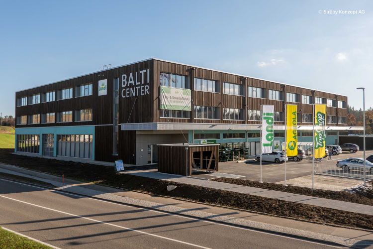 Ein modernes, dreistöckiges Gebäude mit der Aufschrift „BALTI CENTER“ und grünen und gelben Landi-Flaggen davor. Das Gebäude hat große Fenster und Holzvertäfelungen. Im Vordergrund sind ein Parkplatz und eine Straße zu sehen.