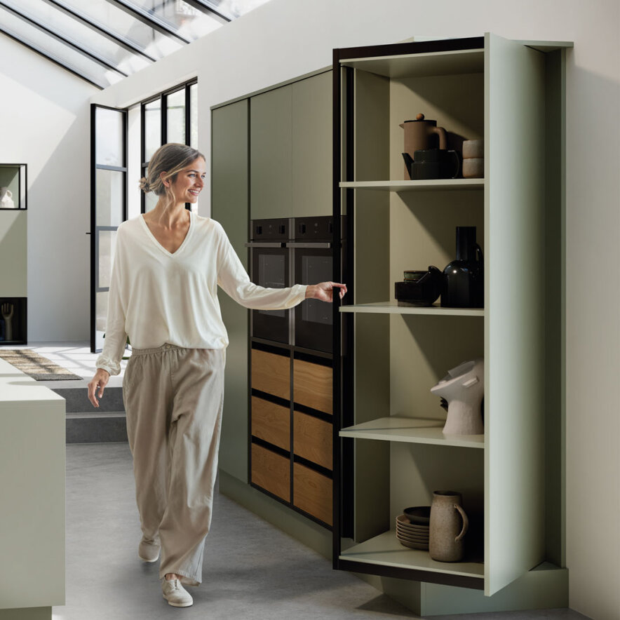 Eine Frau in Freizeitkleidung lächelt, als sie in einer hellen, minimalistischen Küche einen hohen, modernen Küchenschrank mit Regalen für Keramik und Küchenutensilien öffnet.