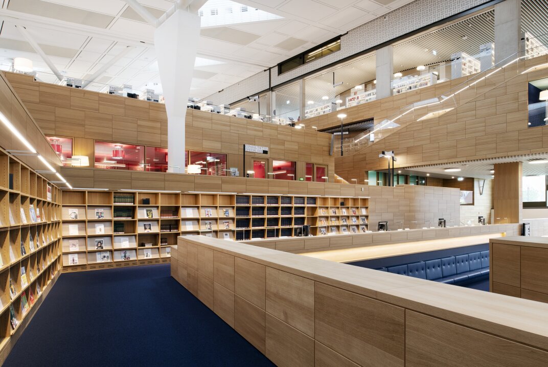 Moderne Bibliothekseinrichtung mit B&uuml;cherregalen aus Holz, blauem Teppich und gro&szlig;z&uuml;gigen Sitzgelegenheiten. Gro&szlig;e Fenster und offene Balkone in den oberen Etagen sorgen f&uuml;r nat&uuml;rliches Licht und den Blick auf weitere B&uuml;cherregale und Arbeitsbereiche.