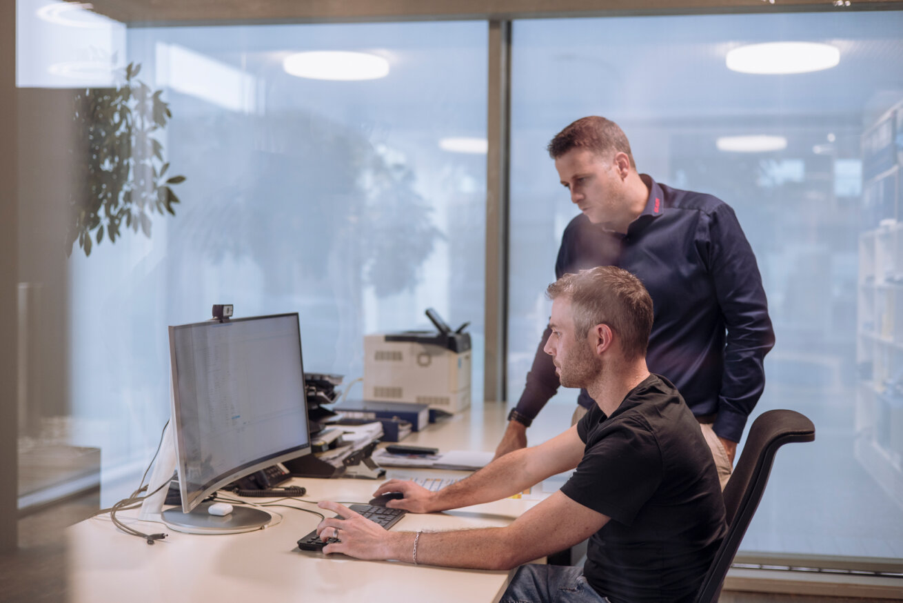 Deux hommes travaillant à un bureau ; l'un est assis et utilise un ordinateur, tandis que l'autre est debout à côté de lui, regardant l'écran. Le bureau est doté de grandes fenêtres et d'un mobilier moderne.