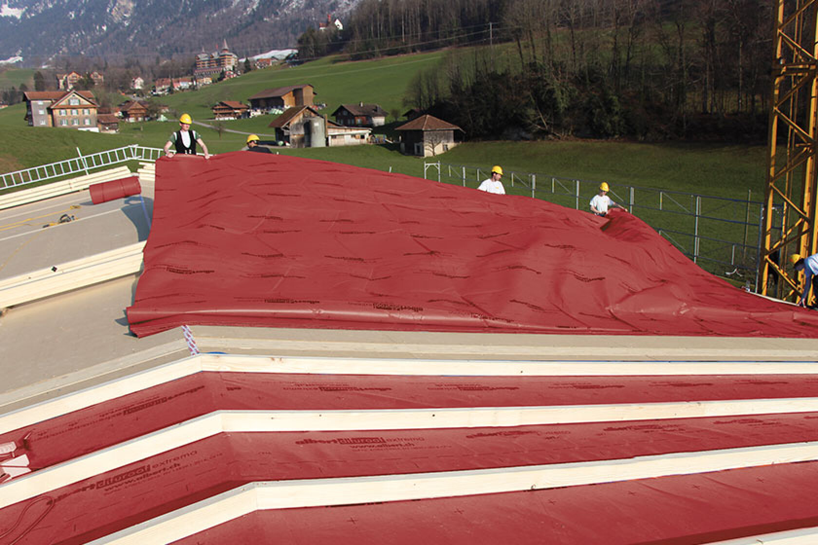 Vier Arbeiter in Schutzkleidung bringen große rote Dämmplatten auf einem Schrägdach an, im Hintergrund ist eine grüne ländliche Landschaft zu sehen, die mit Häusern übersät ist.