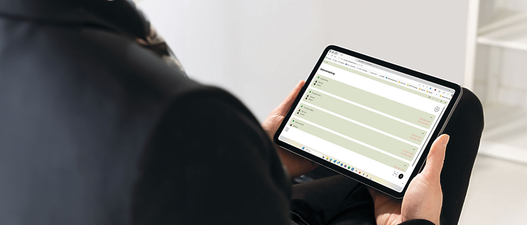 Eine dunkel gekleidete Person hält ein Tablet in der Hand und betrachtet in einem hell beleuchteten Innenraum eine Website mit einer Liste oder einem Tabellenlayout auf dem Bildschirm.