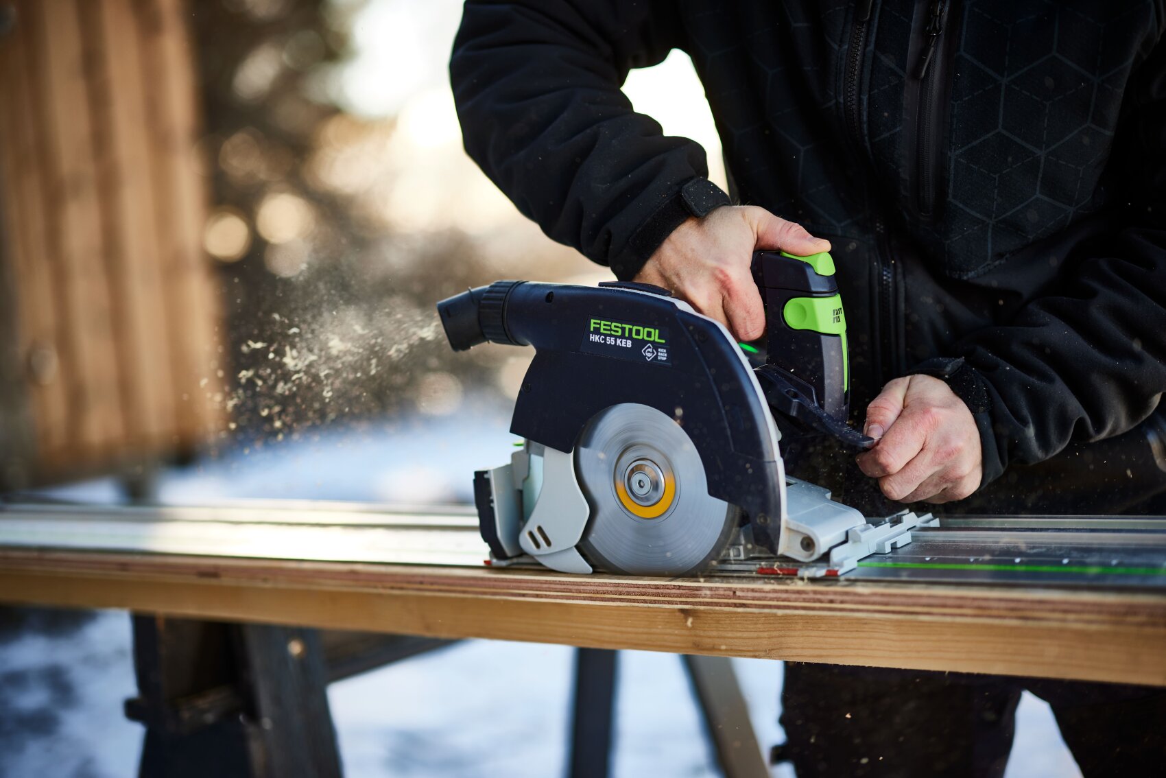 Eine Person in schwarzer Jacke schneidet mit einer Festool-Kreissäge im Freien ein Stück Holz, wobei Sägespäne vom Sägeblatt fliegen.
