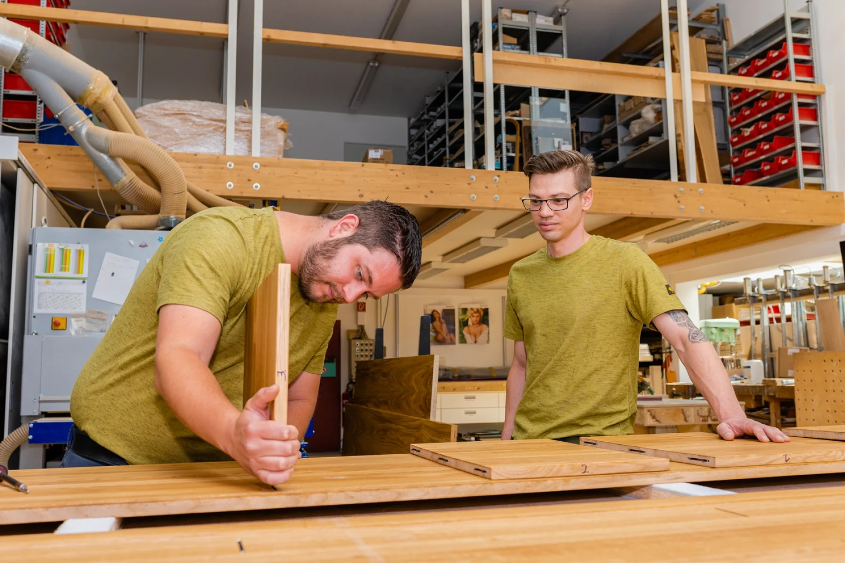 Zwei Männer in passenden grünen Hemden bauen in einer Werkstatt Holzmöbel zusammen. Einer misst ein Brett aus, während der andere beobachtet. Im Hintergrund sind Regale mit Werkzeugen und Materialien zu sehen.