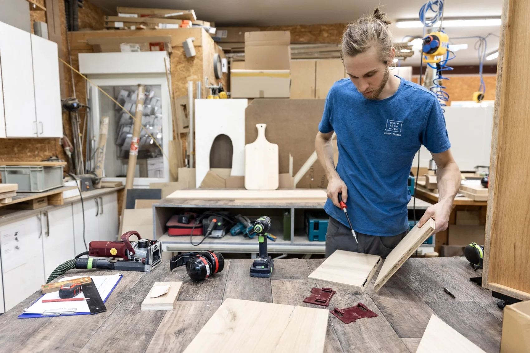 Un homme en chemise bleue travaille à l'assemblage de pièces de bois sur un établi dans un atelier. Des outils, des panneaux de bois et du matériel sont éparpillés autour de lui, et un presse-papiers est posé sur la table. Des étagères et des armoires se trouvent à l'arrière-plan.