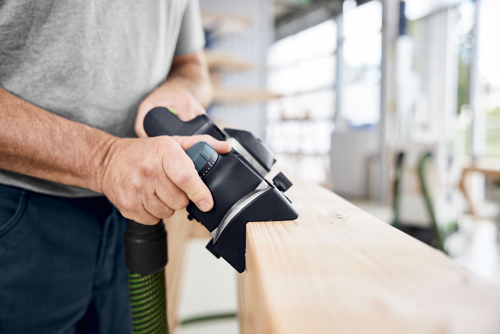 Eine Person glättet in einer gut beleuchteten Werkstatt mit einer elektrischen Hobelmaschine die Oberfläche eines Holzbretts. Nur ihre Hände und das Werkzeug sind sichtbar.