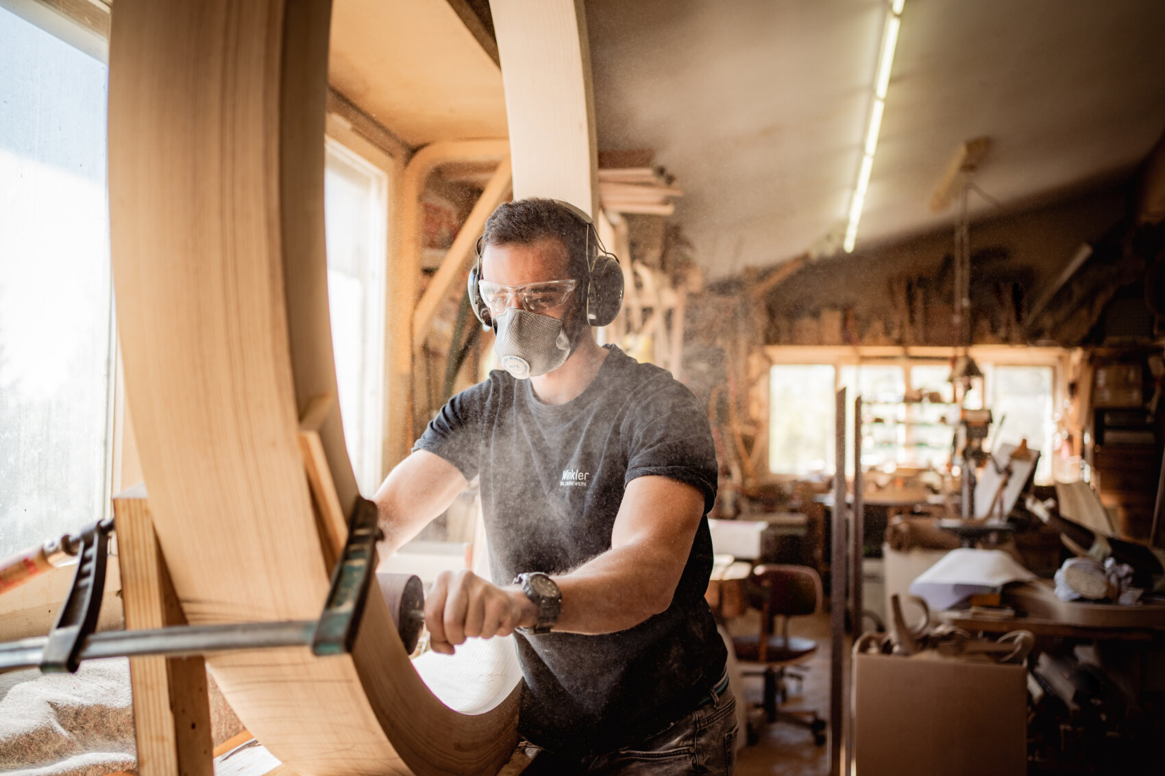 Un homme portant des lunettes de protection, un masque respiratoire et des protections auditives ponce un grand cadre en bois dans un atelier ensoleillé, entouré d'outils de travail du bois et de sciure.