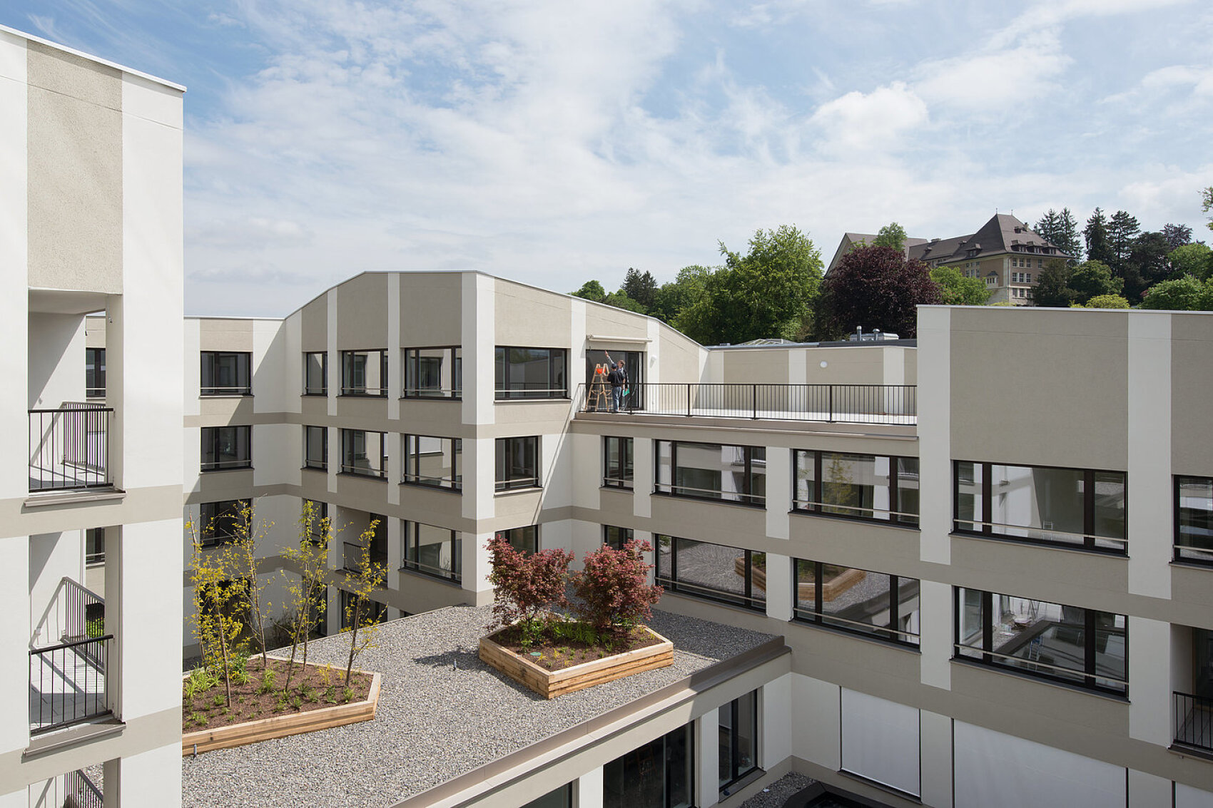 Bâtiment moderne de plusieurs étages avec une cour centrale dotée de parterres de jardin surélevés. Deux personnes se tiennent sur une passerelle supérieure. Des arbres et un bâtiment plus grand sont visibles à l'arrière-plan sous un ciel partiellement nuageux.