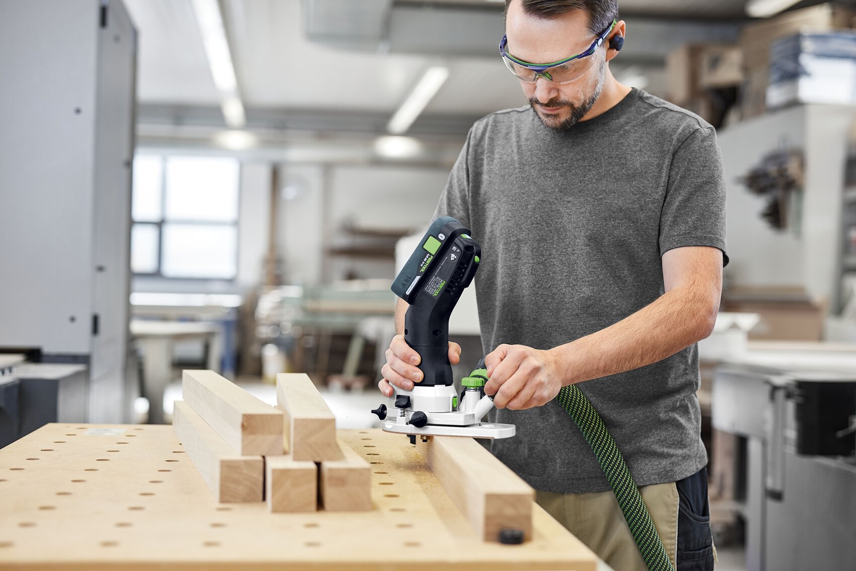 Ein Mann mit Schutzbrille arbeitet mit einem Elektrowerkzeug an einem Holzbrett in einer Werkstatt. Mehrere Holzbretter sind auf einer Werkbank neben ihm gestapelt. Der Arbeitsbereich ist hell und aufgeräumt.