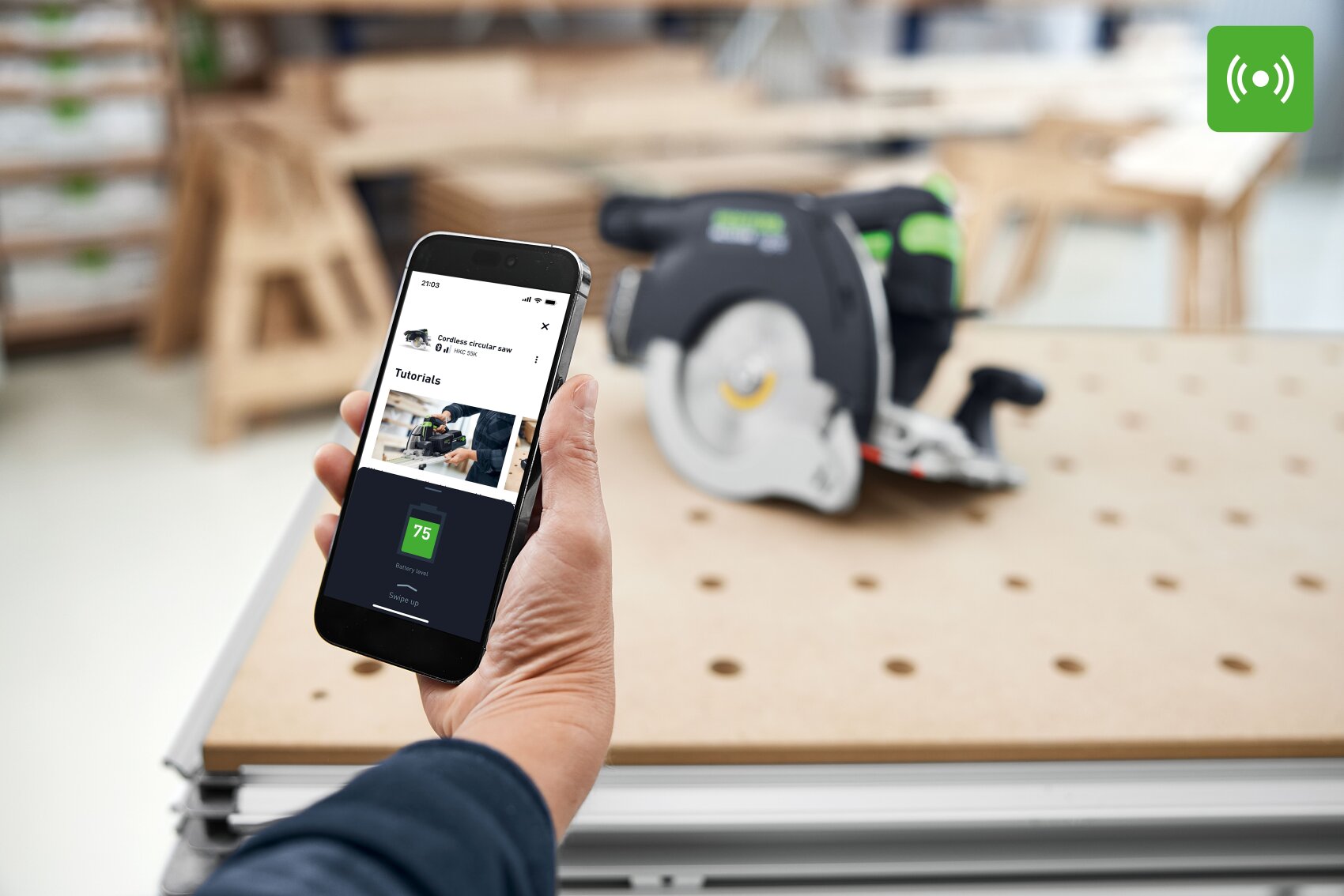 Eine Person mit einem Smartphone, das eine Holzbearbeitungs-App anzeigt, mit einer Kreissäge auf einer perforierten Werkbank im unscharfen Hintergrund; ein grünes Funksignal-Symbol erscheint in der oberen rechten Ecke.