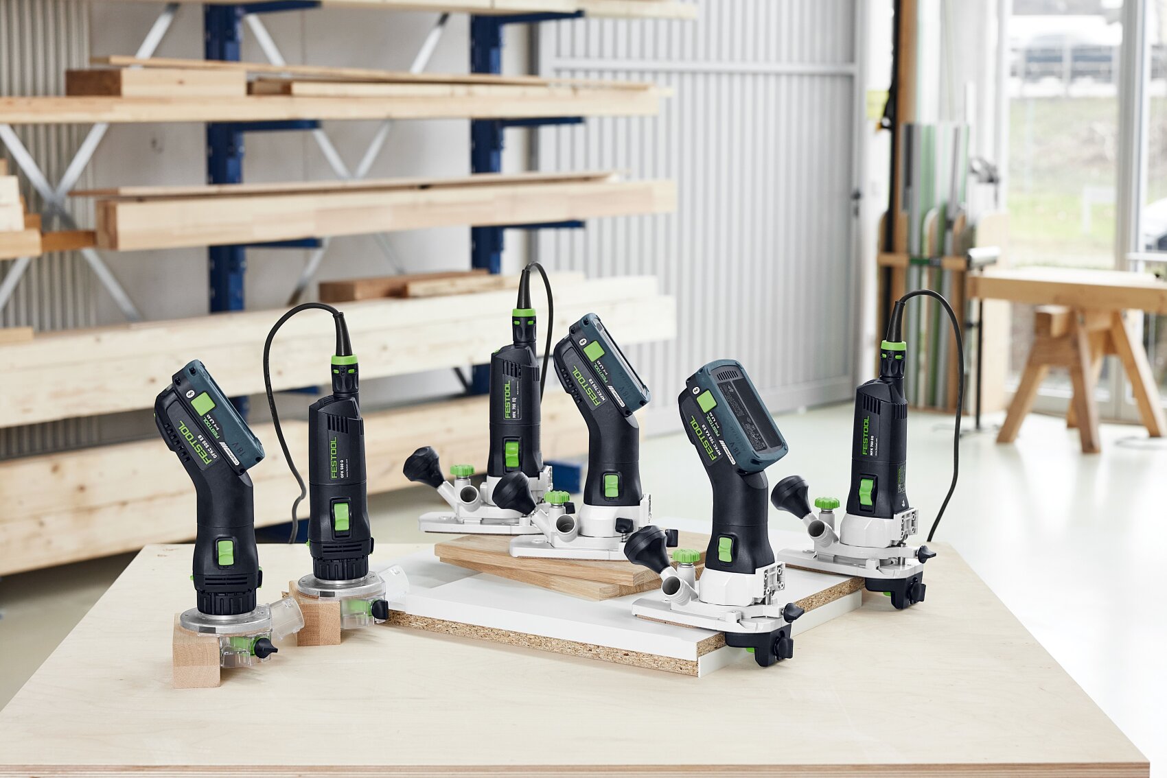 Cinq défonceuses électriques manuelles aux accents noirs et verts sont posées sur un établi dans un atelier de menuiserie clair et moderne, avec des étagères de planches de bois en arrière-plan.