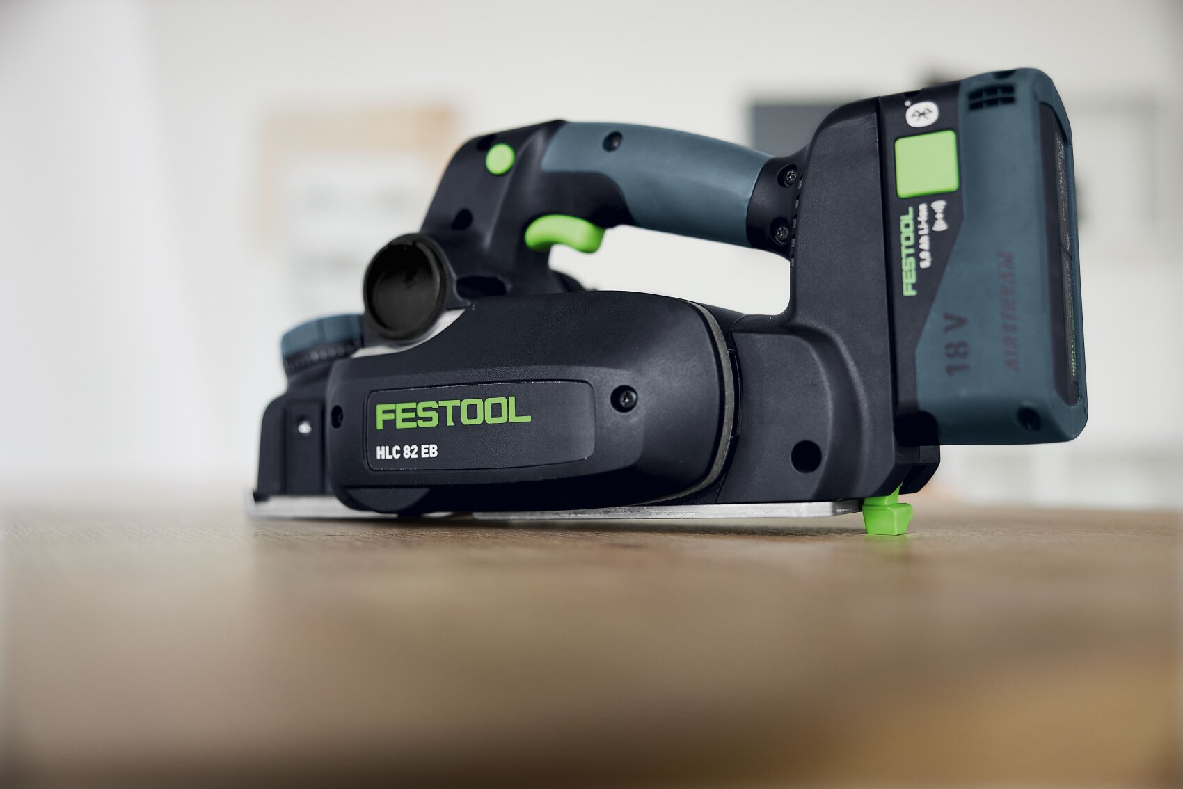 Eine Nahaufnahme eines Festool HLC 82 EB Akku-Handhobels, der auf einer hölzernen Unterlage liegt. Zu sehen sind das schwarz-grüne Design mit sichtbarem Branding und ein angeschlossener 18-V-Akku.