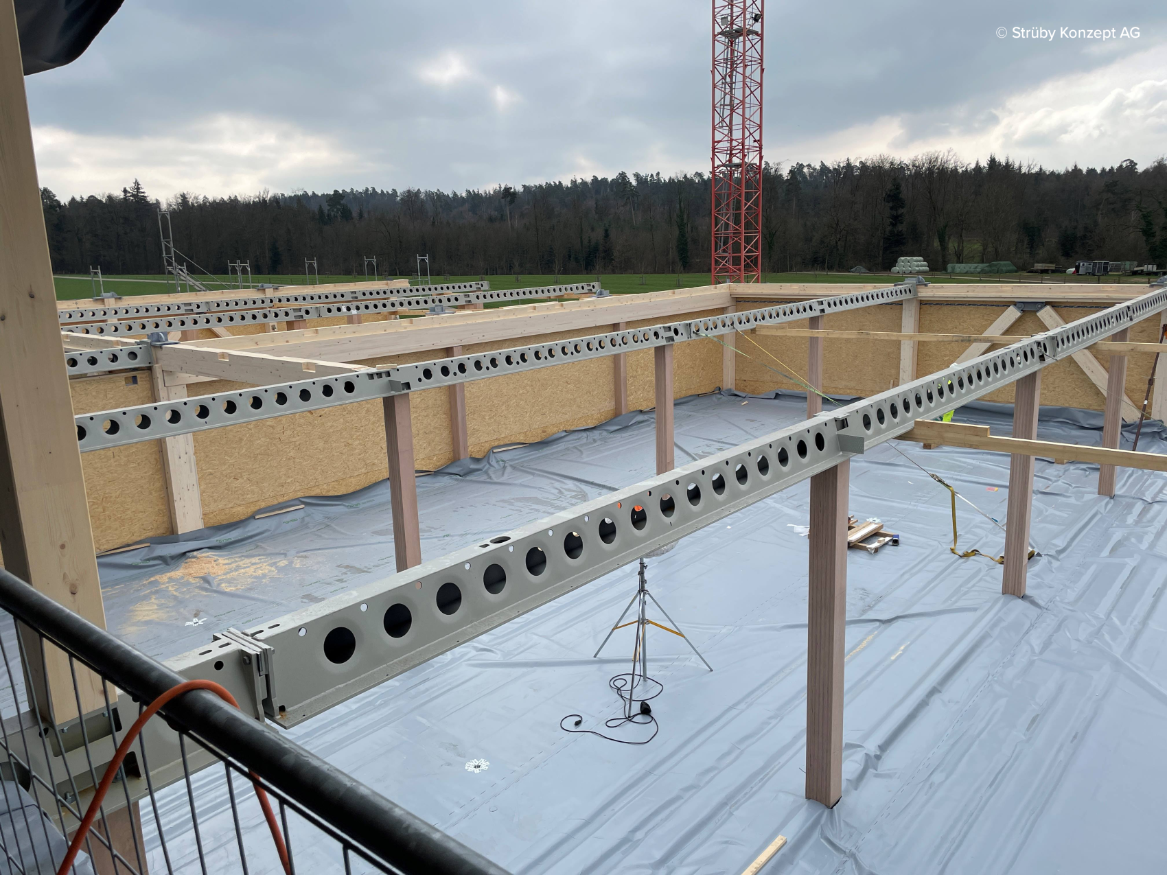Eine Baustelle mit kreisrunden Stahlträgern, Holzwänden und einem kunststoffbeschichteten Boden. Im Hintergrund sind ein roter Kranturm und Bäume unter einem bewölkten Himmel zu sehen.