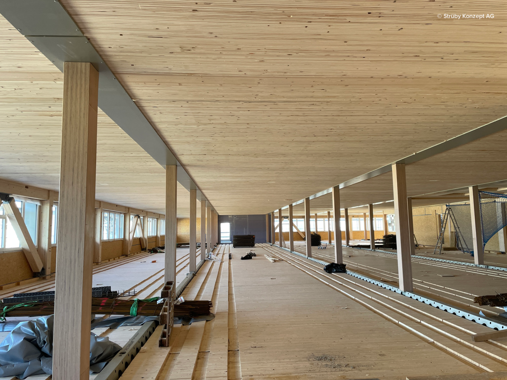Großer, offener Innenraum im Bau mit Holzböden, Holzdecken und Holzsäulen. Tageslicht fällt durch Fensterreihen auf der linken Seite ein. Baumaterialien und Geräte liegen verstreut auf dem Boden.